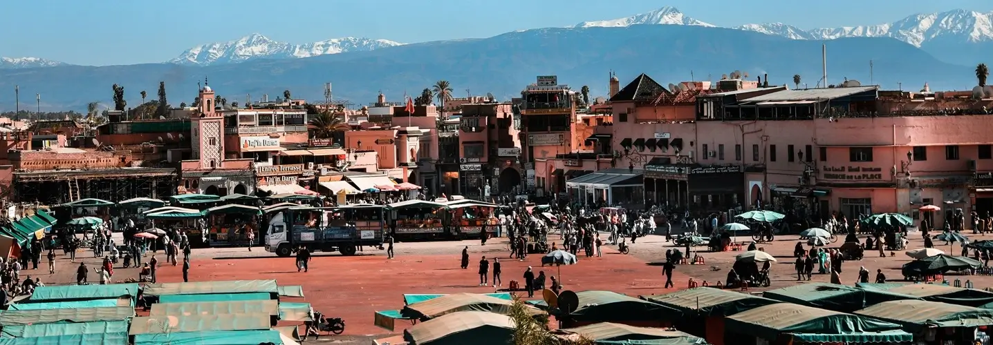 La bulliciosa plaza Jamaa el Fna, con vendedores y multitudes bajo los paraguas, con el Atlas nevado como telón de fondo, como parte de un viaje de 3 días de Marrakech a Fez.